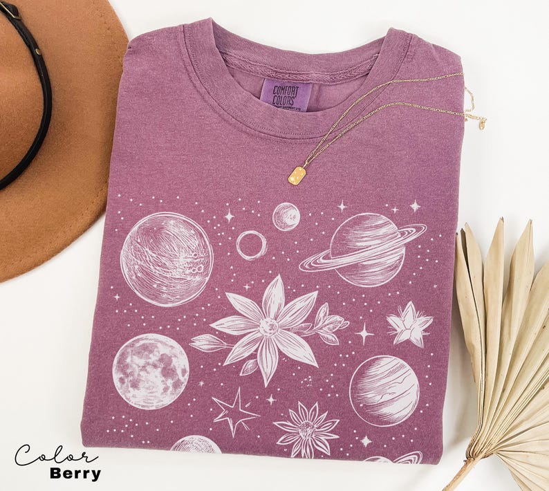 Puede incluir: Una camiseta de color malva con un estampado gr&aacute;fico blanco de planetas, estrellas y flores. El texto "Comfort Colors" est&aacute; impreso en la camiseta.