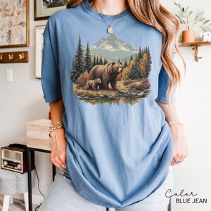 May include: A light blue, short-sleeved t-shirt featuring a detailed illustration of a bear and cub in a forest setting with a mountain backdrop. The text "Color BLUE JEAN" is printed in a script font at the bottom right.