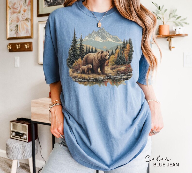 May include: A light blue, short-sleeved t-shirt featuring a detailed illustration of a bear and cub in a forest setting with a mountain backdrop. The text "Color BLUE JEAN" is printed in a script font at the bottom right.