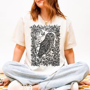 May include: Ivory-colored t-shirt featuring a detailed black and white illustration of an owl within a floral frame. The design includes a butterfly and botanical elements. The shirt is a relaxed fit.