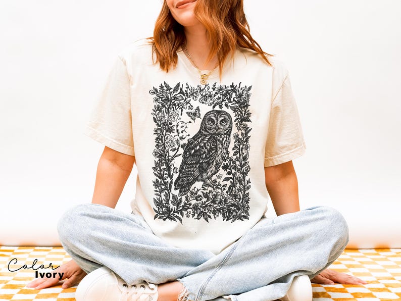 May include: Ivory-colored t-shirt featuring a detailed black and white illustration of an owl within a floral frame. The design includes a butterfly and botanical elements. The shirt is a relaxed fit.
