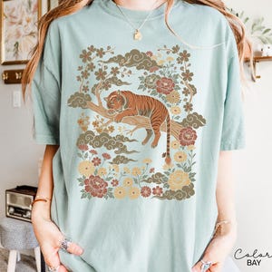 May include: A light blue t-shirt featuring a detailed illustration of a tiger resting on a tree branch, surrounded by flowers and clouds. The tiger is brown with black stripes. The design has a vintage, Asian-inspired aesthetic.