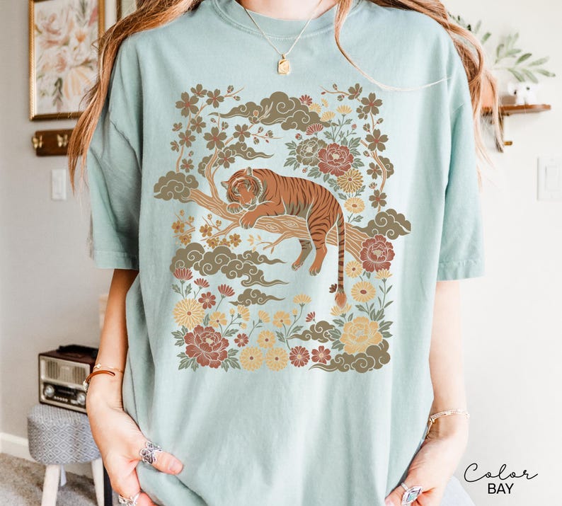 May include: A light blue t-shirt featuring a detailed illustration of a tiger resting on a tree branch, surrounded by flowers and clouds. The tiger is brown with black stripes. The design has a vintage, Asian-inspired aesthetic.