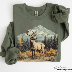 May include: An olive green sweatshirt featuring a detailed graphic of a deer with large antlers standing in front of a mountain range and evergreen trees. The text "Color Military Green" is at the bottom right.