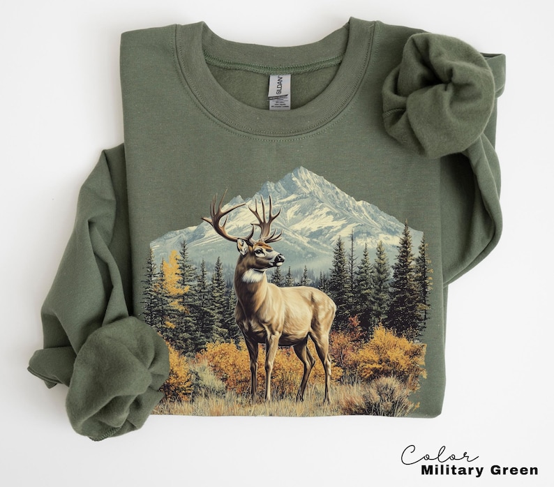 May include: An olive green sweatshirt featuring a detailed graphic of a deer with large antlers standing in front of a mountain range and evergreen trees. The text "Color Military Green" is at the bottom right.
