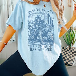May include: Light blue t-shirt with a graphic featuring a knight riding a tiger, a castle, and the text "HEAR YE! HEAR YE!" and "THE FUN AUNT HAS ARRIVED". The shirt is a solid color and has short sleeves.
