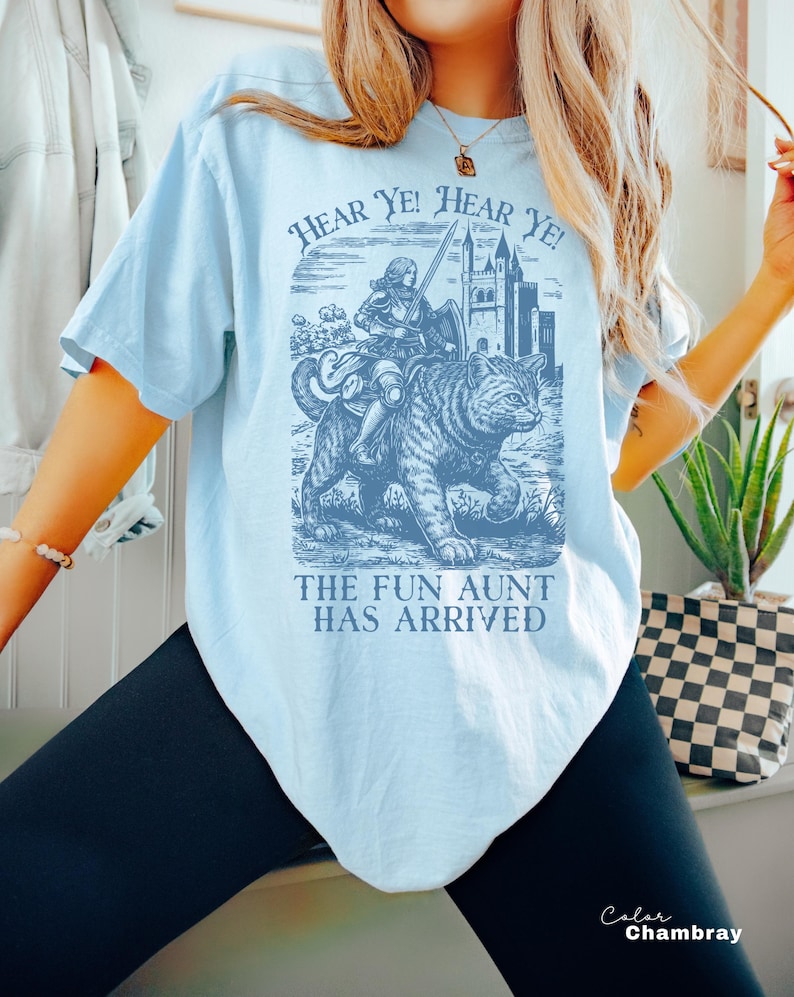 May include: Light blue t-shirt with a graphic featuring a knight riding a tiger, a castle, and the text "HEAR YE! HEAR YE!" and "THE FUN AUNT HAS ARRIVED". The shirt is a solid color and has short sleeves.