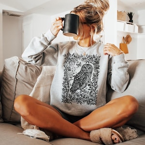 May include: A light gray sweatshirt featuring a detailed black and white owl illustration within a floral frame. The sweatshirt is labeled "Color Ash" and is being worn.