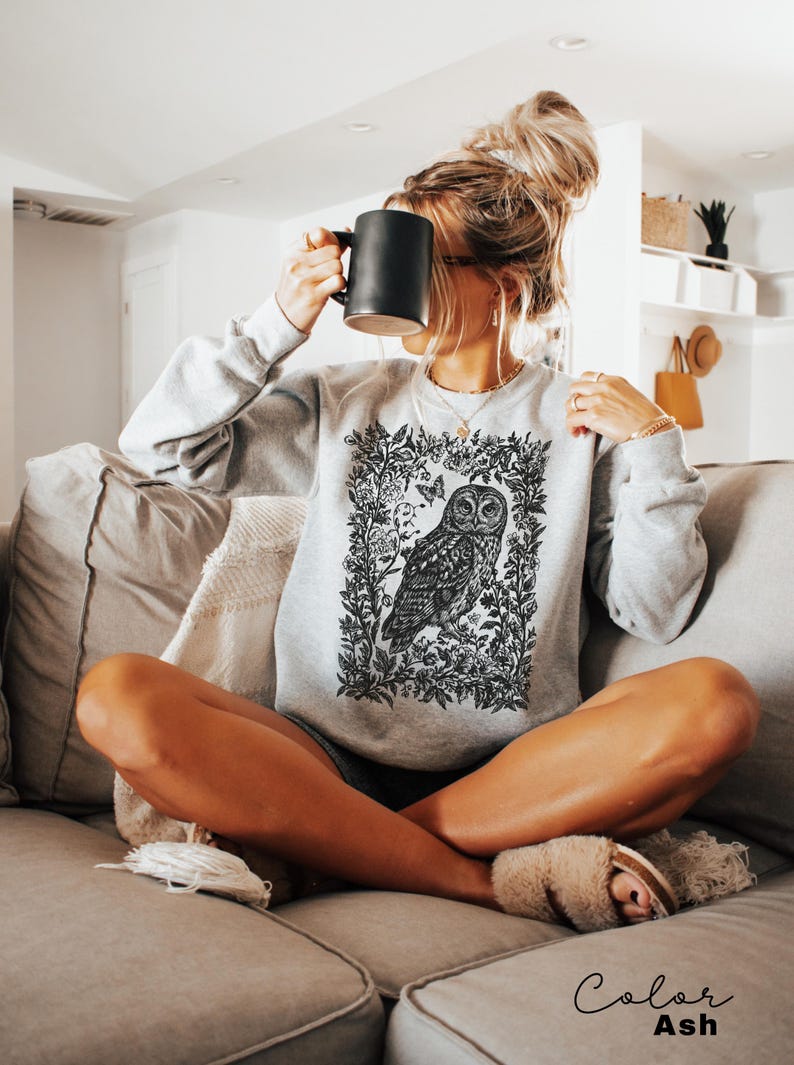 May include: A light gray sweatshirt featuring a detailed black and white owl illustration within a floral frame. The sweatshirt is labeled "Color Ash" and is being worn.