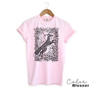 Pu&ograve; includere: T-shirt rosa chiaro con un'illustrazione in bianco e nero di una volpe che salta all'interno di una cornice floreale. Il testo "Color Blossom" &egrave; scritto in corsivo in basso a destra. La maglietta &egrave; appesa a una gruccia di legno.