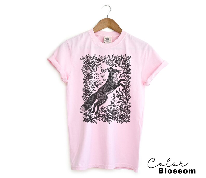 Pu&ograve; includere: T-shirt rosa chiaro con un'illustrazione in bianco e nero di una volpe che salta all'interno di una cornice floreale. Il testo "Color Blossom" &egrave; scritto in corsivo in basso a destra. La maglietta &egrave; appesa a una gruccia di legno.