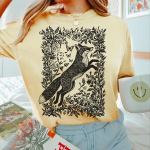Pu&ograve; includere: Una felpa giallo chiaro con un'illustrazione in bianco e nero di una volpe che salta all'interno di una cornice floreale, con farfalle. Il testo "Color Banana" &egrave; in basso a destra.