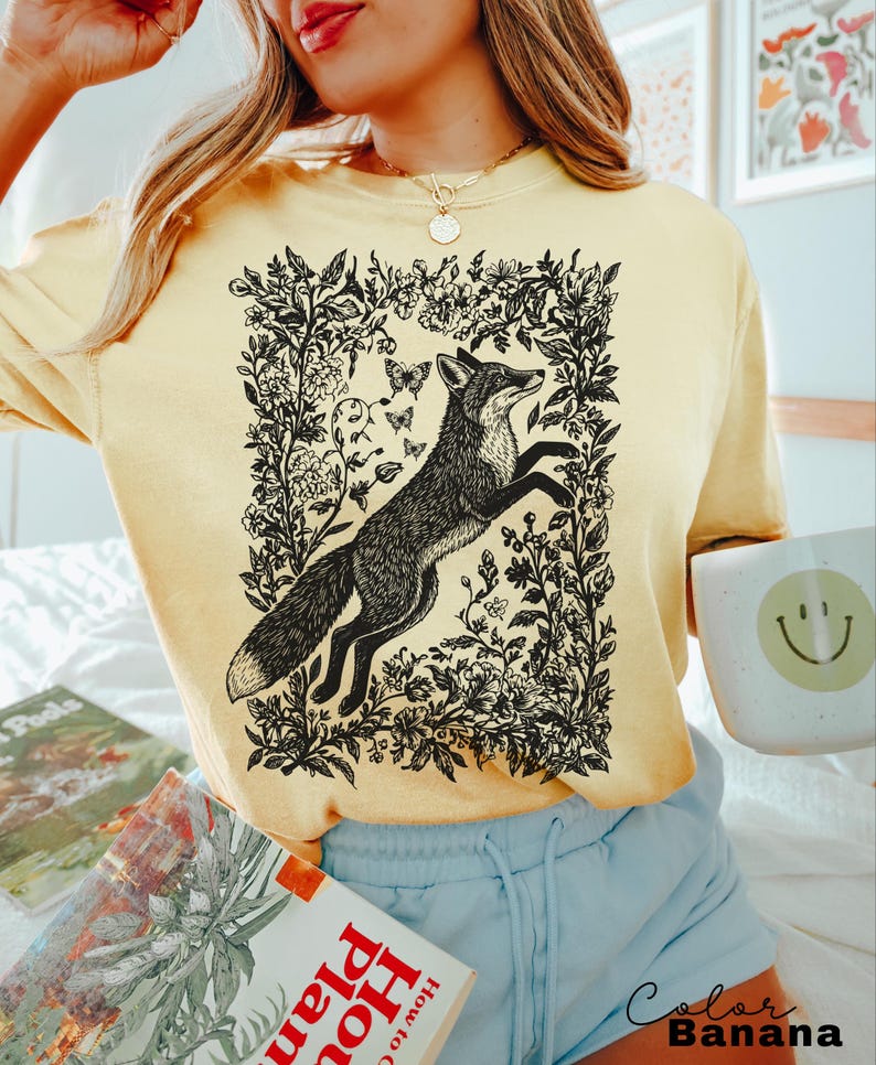 Pu&ograve; includere: Una felpa giallo chiaro con un'illustrazione in bianco e nero di una volpe che salta all'interno di una cornice floreale, con farfalle. Il testo "Color Banana" &egrave; in basso a destra.