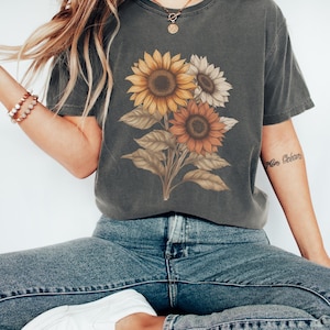 May include: A grey t-shirt featuring a graphic of three sunflowers in shades of yellow, orange, and white. The sunflowers are arranged in a bouquet with green leaves.