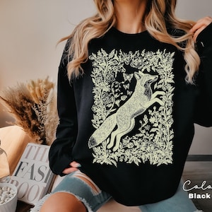 Pu&ograve; includere: Felpa nera con un'illustrazione color crema di una volpe che salta tra elementi floreali e farfalle. Il disegno &egrave; incorniciato da un bordo di fiori e foglie. Il testo "Color Black" &egrave; visibile in basso a destra.