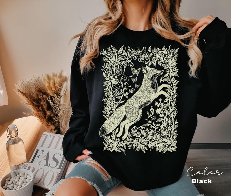 Pu&ograve; includere: Felpa nera con un'illustrazione color crema di una volpe che salta tra elementi floreali e farfalle. Il disegno &egrave; incorniciato da un bordo di fiori e foglie. Il testo "Color Black" &egrave; visibile in basso a destra.