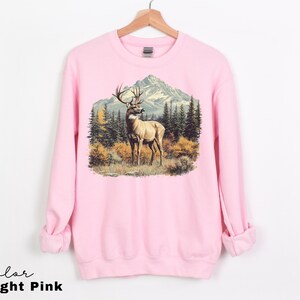 May include: Light pink sweatshirt featuring a detailed graphic of a deer in a forest scene with mountains in the background. The deer has large antlers and is standing in a field of grass and bushes. The text "Color Light Pink" is at the bottom.
