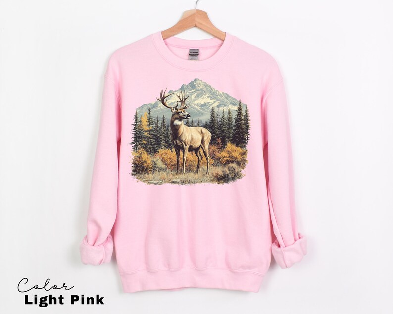 May include: Light pink sweatshirt featuring a detailed graphic of a deer in a forest scene with mountains in the background. The deer has large antlers and is standing in a field of grass and bushes. The text "Color Light Pink" is at the bottom.