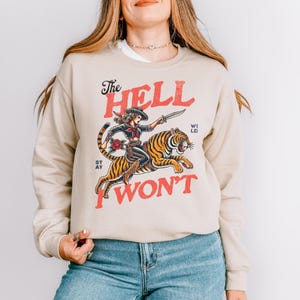 May include: Beige sweatshirt with a graphic of a woman riding a tiger, holding a knife. The text "The Hell I Won't" is in red. The woman has a red scarf and a black hat. The tiger is orange and black.