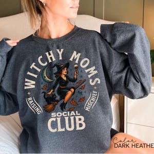 May include: Dark heather grey sweatshirt with a graphic that reads "Witchy Moms Social Club." The design features a witch on a broom, a black cat, a crescent moon, and autumn leaves. The text "Raising Mischief" is also included.