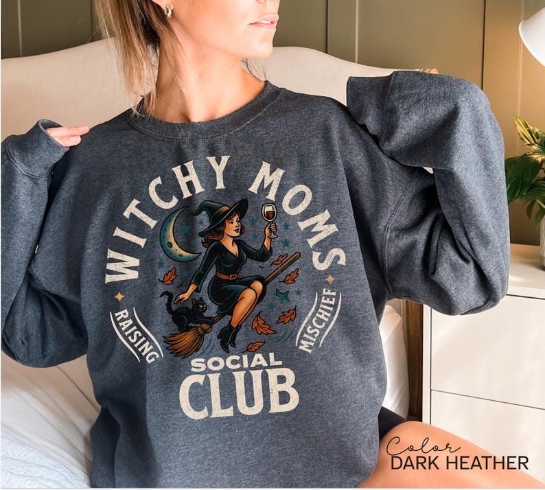 May include: Dark heather grey sweatshirt with a graphic that reads "Witchy Moms Social Club." The design features a witch on a broom, a black cat, a crescent moon, and autumn leaves. The text "Raising Mischief" is also included.
