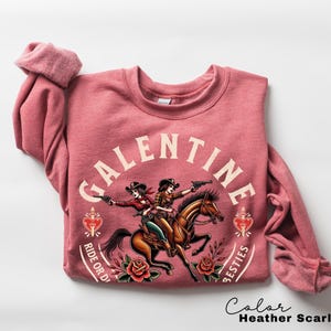 May include: Heather scarlet red sweatshirt with a graphic of two cowgirls on horseback, guns drawn. The word "Galentine" arches above the image, with "Ride or Die" and "Besties" below. Roses and hearts complete the design.