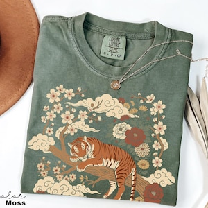 May include: Moss green t-shirt featuring a tiger resting on a tree branch, surrounded by floral and cloud designs. The shirt has a crew neck and short sleeves. A silver necklace with a round pendant is on top of the shirt.