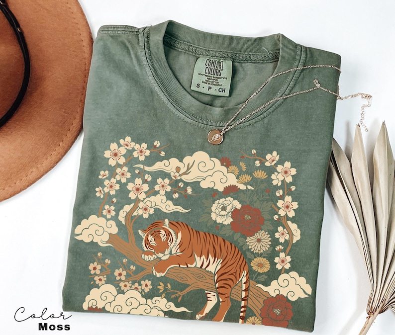 May include: Moss green t-shirt featuring a tiger resting on a tree branch, surrounded by floral and cloud designs. The shirt has a crew neck and short sleeves. A silver necklace with a round pendant is on top of the shirt.