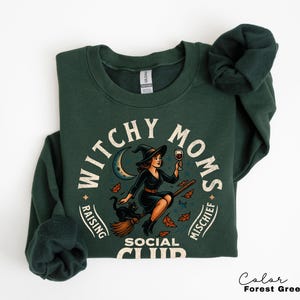 May include: Forest green sweatshirt with a graphic that reads "Witchy Moms Social Club". The design features a witch on a broom holding a glass of wine, with a black cat and crescent moon.