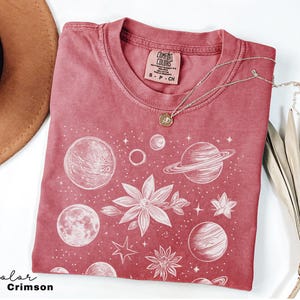 Puede incluir: Una camiseta de color rojo carmes&iacute; con un estampado gr&aacute;fico blanco de planetas, estrellas y flores. La camiseta tiene un aspecto desgastado y el texto "Comfort Colors" est&aacute; impreso en la etiqueta.
