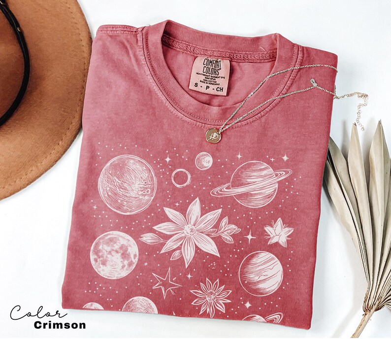 Puede incluir: Una camiseta de color rojo carmes&iacute; con un estampado gr&aacute;fico blanco de planetas, estrellas y flores. La camiseta tiene un aspecto desgastado y el texto "Comfort Colors" est&aacute; impreso en la etiqueta.