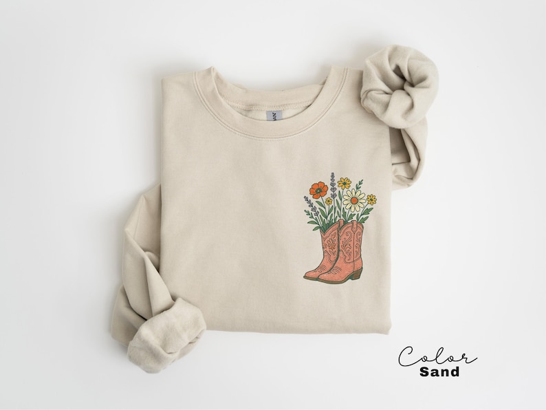 Puede incluir: Sudadera color arena con un gr&aacute;fico de botas de vaquero florales. El gr&aacute;fico presenta un par de botas de vaquero de color coral llenas de flores naranjas, amarillas y moradas. El texto "Color Sand" est&aacute; escrito en una fuente cursiva.
