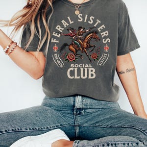 May include: A charcoal grey t-shirt with a vintage-style graphic of two women riding a horse. The graphic includes the text "Feral Sisters Social Club" and "Ride or Die".