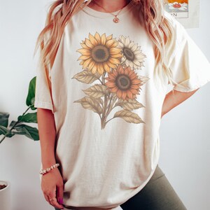 May include: A cream coloured t-shirt with a graphic of three sunflowers in shades of yellow, orange, and brown. The text "Color Ivory" is at the bottom of the image.
