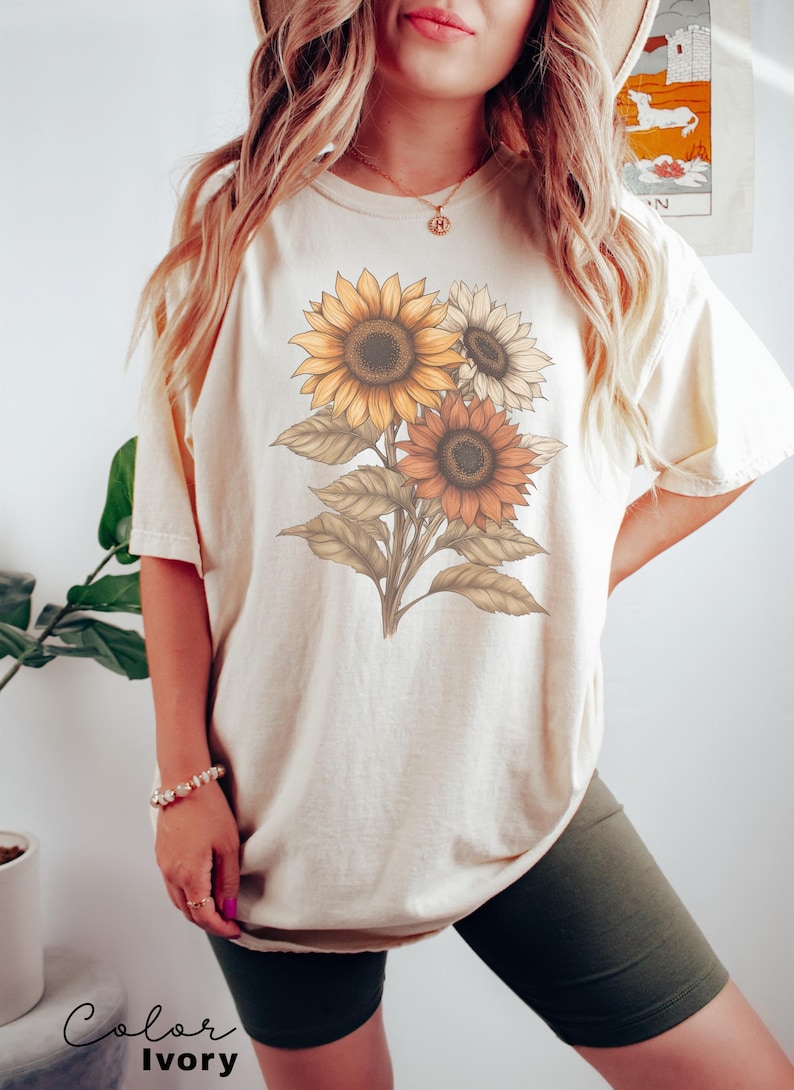 May include: A cream coloured t-shirt with a graphic of three sunflowers in shades of yellow, orange, and brown. The text "Color Ivory" is at the bottom of the image.