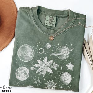 Puede incluir: Una camiseta verde con un estampado blanco de planetas, estrellas y flores. El texto "COMFORT COLORS" y "S - P - CH" est&aacute; impreso en la etiqueta.