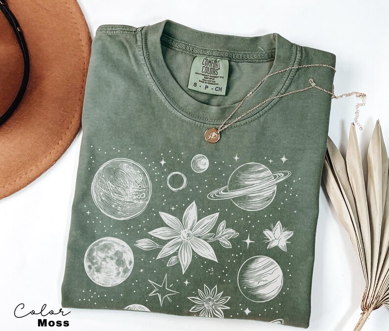 Puede incluir: Una camiseta verde con un estampado blanco de planetas, estrellas y flores. El texto "COMFORT COLORS" y "S - P - CH" est&aacute; impreso en la etiqueta.