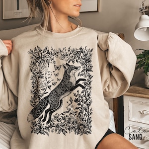 Pu&ograve; includere: Felpa color sabbia con un'illustrazione in bianco e nero di una volpe che salta all'interno di una cornice floreale, con farfalle. La felpa ha maniche lunghe e girocollo. Il colore &egrave; etichettato "SAND" in basso a destra.