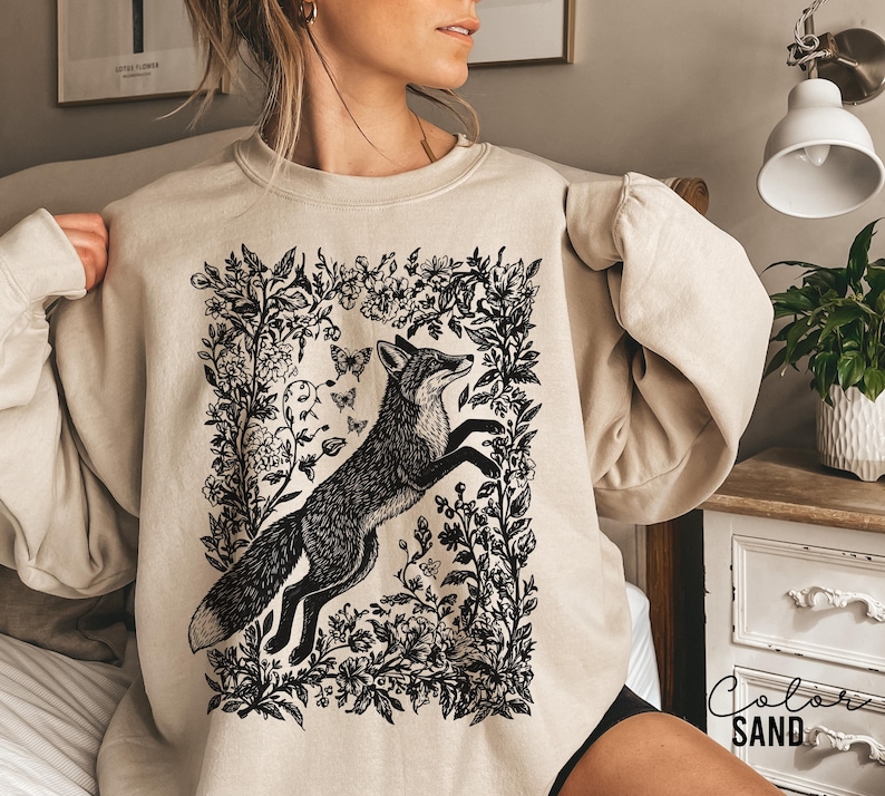 Pu&ograve; includere: Felpa color sabbia con un'illustrazione in bianco e nero di una volpe che salta all'interno di una cornice floreale, con farfalle. La felpa ha maniche lunghe e girocollo. Il colore &egrave; etichettato "SAND" in basso a destra.
