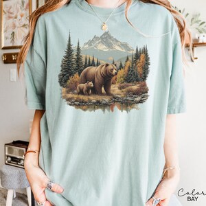 May include: A light blue t-shirt featuring a detailed illustration of a brown bear and a cub in a forest setting with a mountain backdrop. The design includes trees, a lake, and autumn colors.
