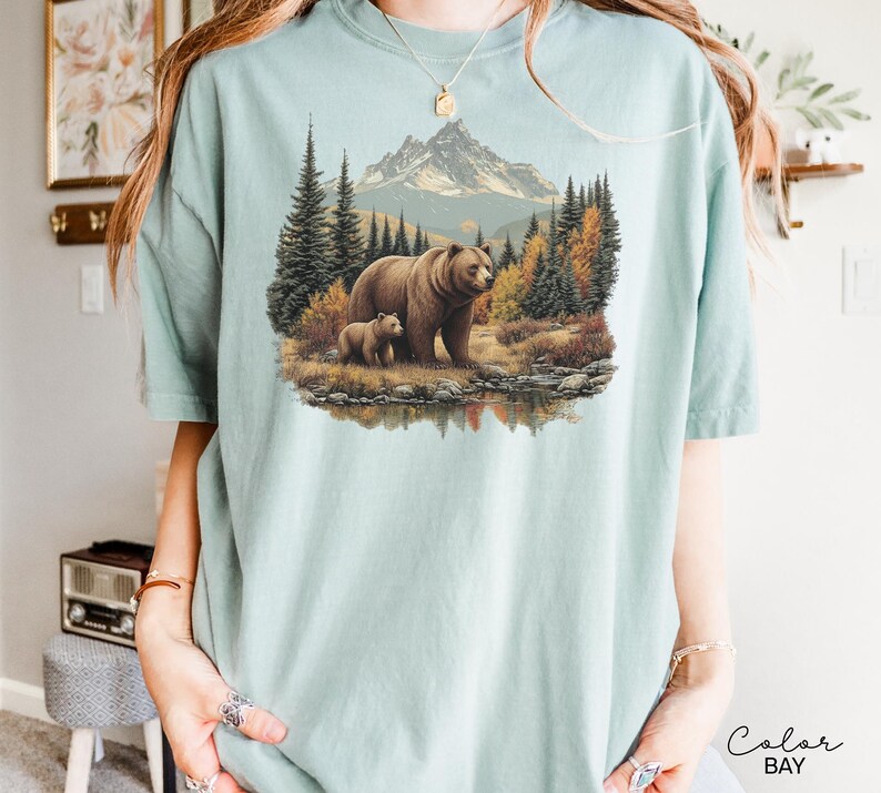 May include: A light blue t-shirt featuring a detailed illustration of a brown bear and a cub in a forest setting with a mountain backdrop. The design includes trees, a lake, and autumn colors.