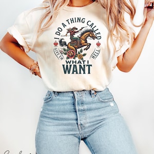 May include: Ivory t-shirt featuring a graphic of a woman riding a horse with the text "I do a thing called give 'em what I want".