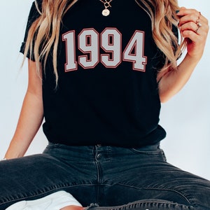 May include: Black t-shirt with a white 1994 graphic. The shirt is worn by a person with long blonde hair and is sitting on a red chair.