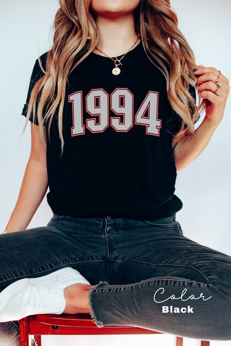 May include: Black t-shirt with a white 1994 graphic. The shirt is worn by a person with long blonde hair and is sitting on a red chair.