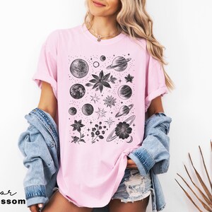 Puede incluir: Una camiseta rosa con un estampado gr&aacute;fico en blanco y negro de planetas, estrellas y flores. El gr&aacute;fico est&aacute; centrado en la camiseta.