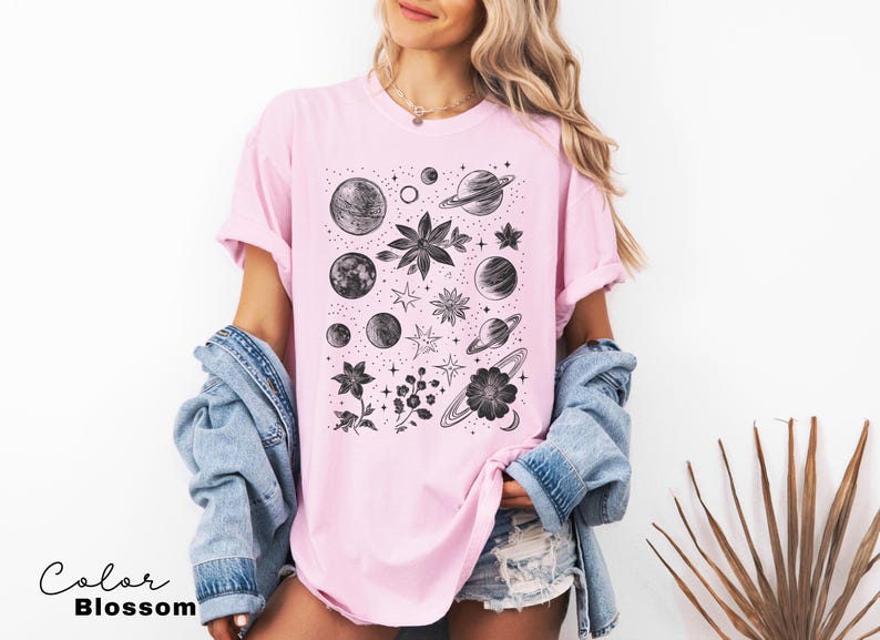 Puede incluir: Una camiseta rosa con un estampado gr&aacute;fico en blanco y negro de planetas, estrellas y flores. El gr&aacute;fico est&aacute; centrado en la camiseta.