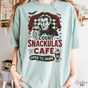 May include: A light blue t-shirt featuring a graphic of Count Dracula holding a milkshake, with the text "Count Snackula's Cafe" in maroon. The design includes bats and a checkered curtain. The shirt also says "Open Til Dawn".