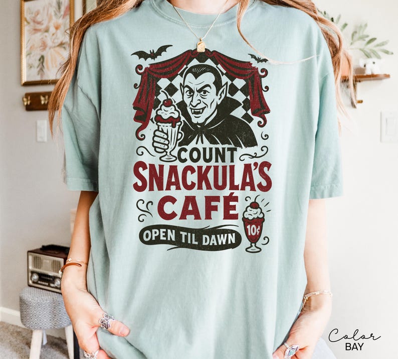May include: A light blue t-shirt featuring a graphic of Count Dracula holding a milkshake, with the text "Count Snackula's Cafe" in maroon. The design includes bats and a checkered curtain. The shirt also says "Open Til Dawn".