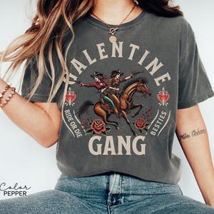 May include: Dark gray t-shirt with a graphic of two cowgirls riding a horse, guns drawn, surrounded by roses and hearts. The text reads "VALENTINE GANG, RIDE OR DIE, BESTIES".