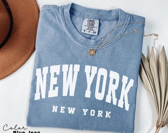 May include: A blue jean colored t-shirt with the words "NEW YORK" in large white block letters. The words "NEW YORK" are also printed in smaller white letters below. A gold necklace with a pendant is draped over the shirt.
